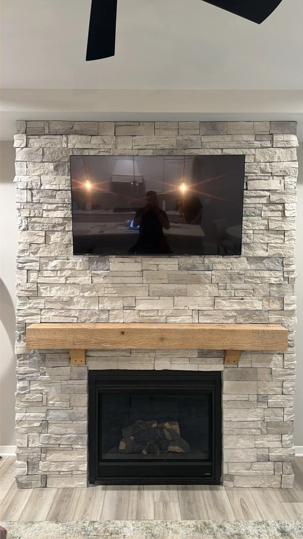 TV above a wood mantel on a light stacked stone fireplace